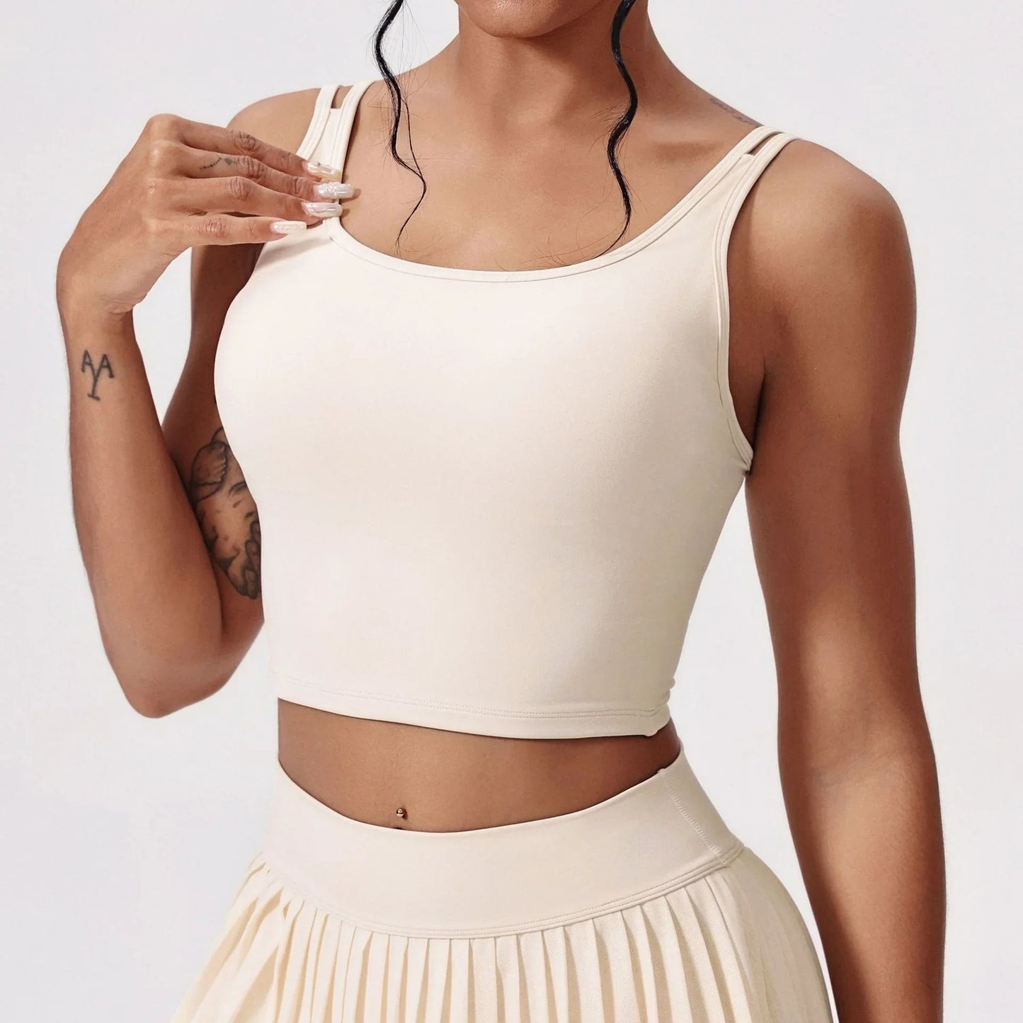Woman wearing beige double strap crop top and pleated skirt with tattoos and belly piercing