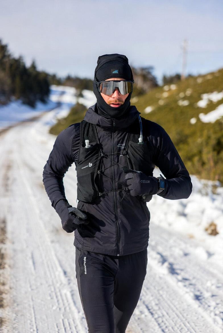Man running in snow wearing black winter running gear, hydration vest, and reflective goggles