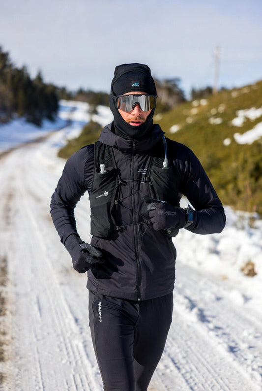 Man running in snow wearing black winter running gear, hydration vest, and reflective goggles